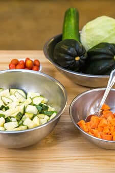 Silver mixing bowls hold chopped zucchini, chopped carrots, and acorn squash