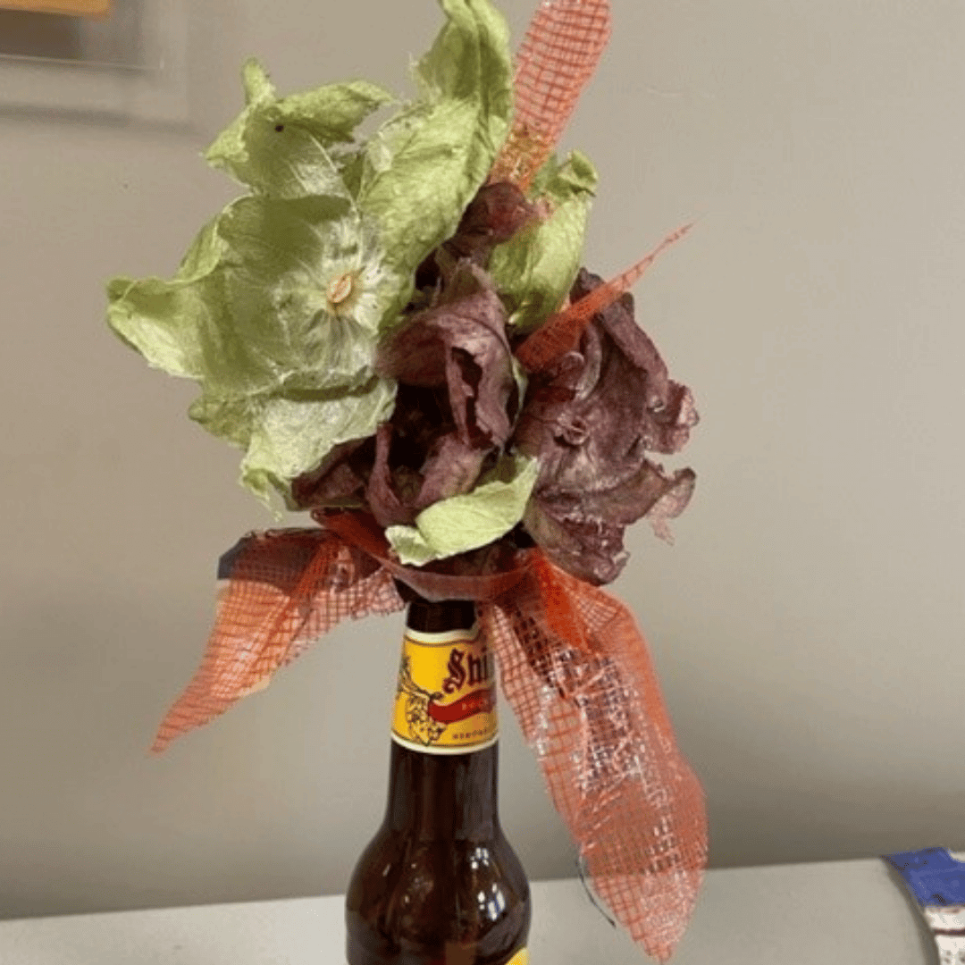 Flower arrangement made from tomatillo husks died in beet water, an orange bag, and beer bottle