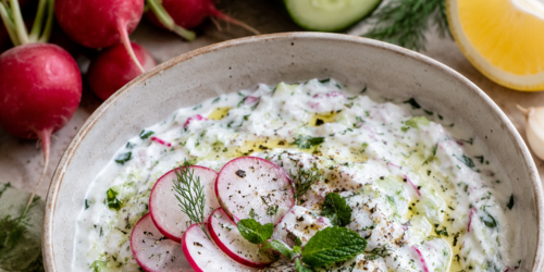 A bowl filled with radish and cucumber Tzatziki on a table with fresh radishes, cucumber, and a lemon wedge surrounding it.